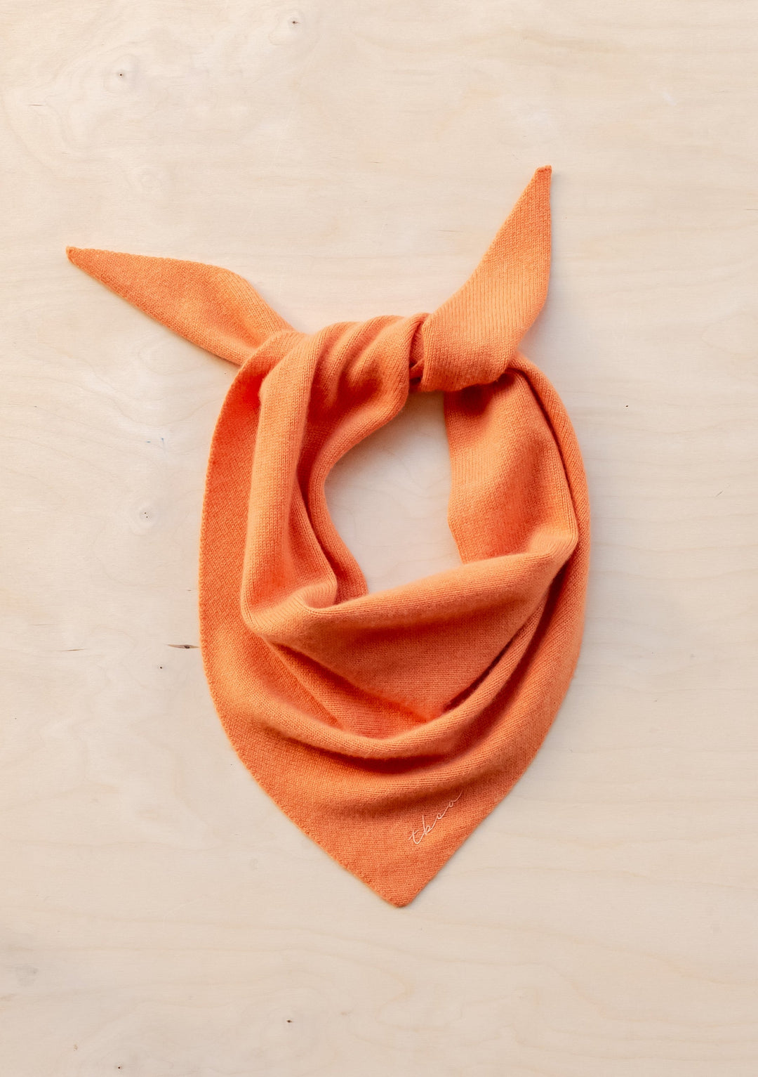 Merino Triangle Scarf in Apricot elegantly tied in a classic bandana style, showcasing its soft wool texture and warm apricot color against a light beige background. The triangular shape creates graceful folds and draping, highlighting the scarf's versatile styling potential.