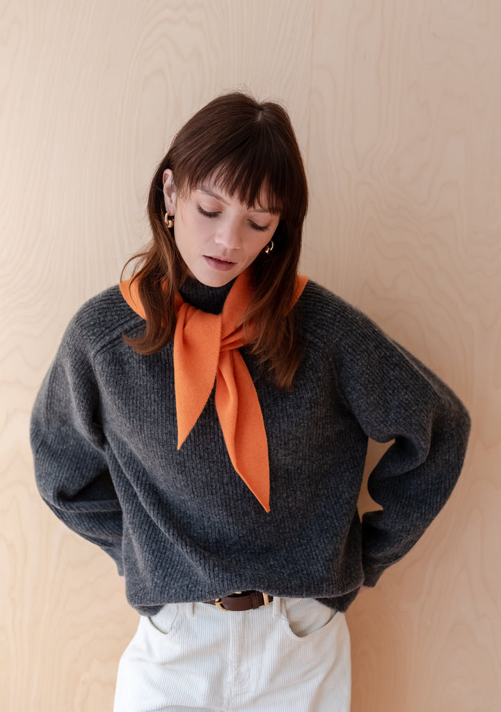 Merino Triangle Scarf in Apricot styled on a model wearing a chunky gray sweater and white pants, showcasing the vibrant apricot-colored scarf tied loosely around the neck against a light wooden background. The scarf's soft merino wool texture and casual draping demonstrate its versatile styling potential.