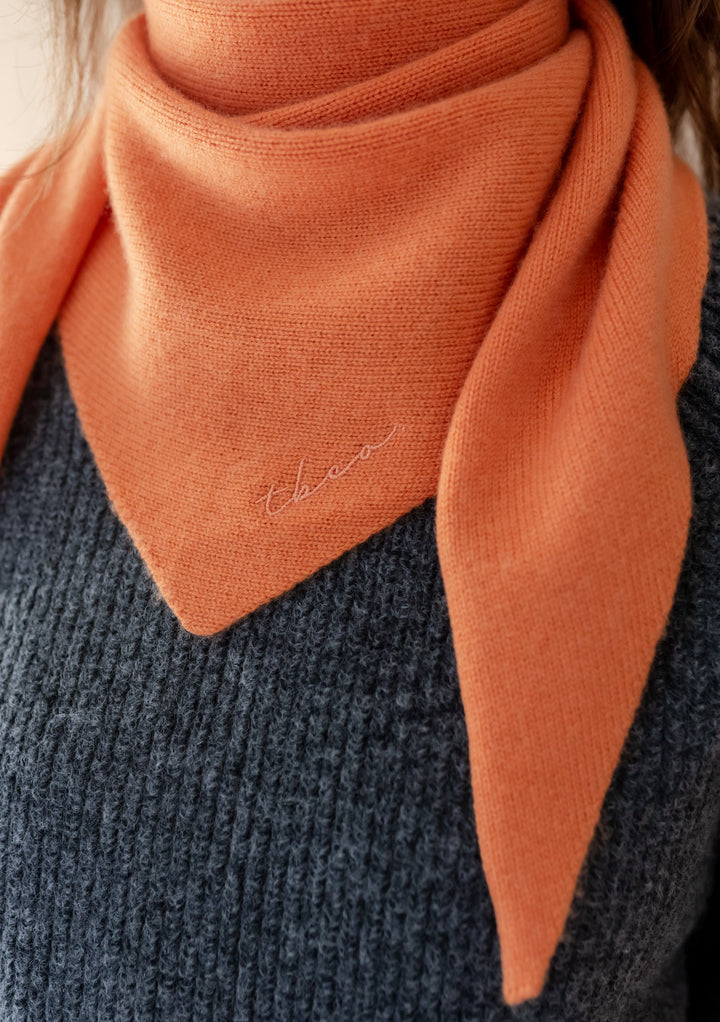 Merino Triangle Scarf in Apricot draped elegantly around neck, showing soft knit texture against dark gray sweater. Small embroidered logo visible on corner of the triangular scarf. Warm apricot color highlights the natural drape and versatility of the merino wool accessory.