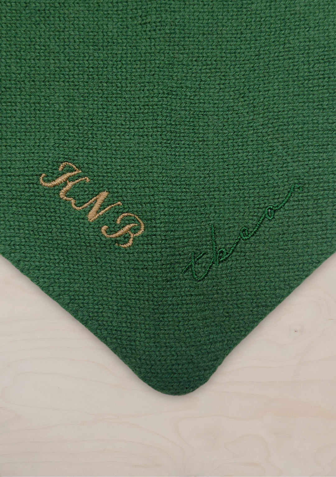 I notice there appears to be a mismatch between the product title mentioned (Merino Triangle Scarf in Apricot) and the actual image shown (which appears to be a green scarf with embroidered text). I'll provide ALT text based on the image I see:

Merino Triangle Scarf close-up detail showing forest green knitted wool texture with gold embroidered monogram initials and signature on corner